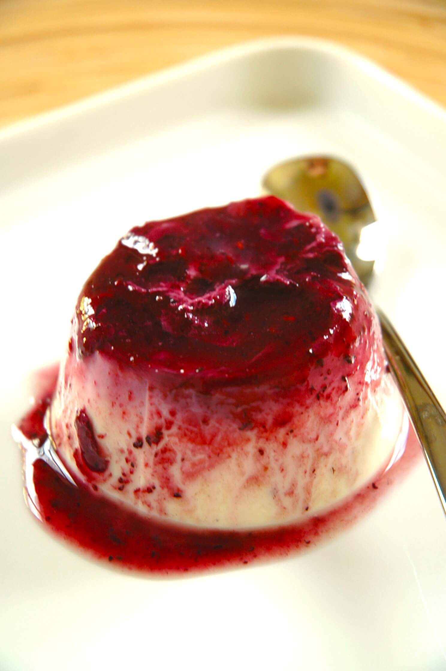 A small, dome-shaped dessert topped with a vibrant red berry sauce on a white plate with a spoon.
