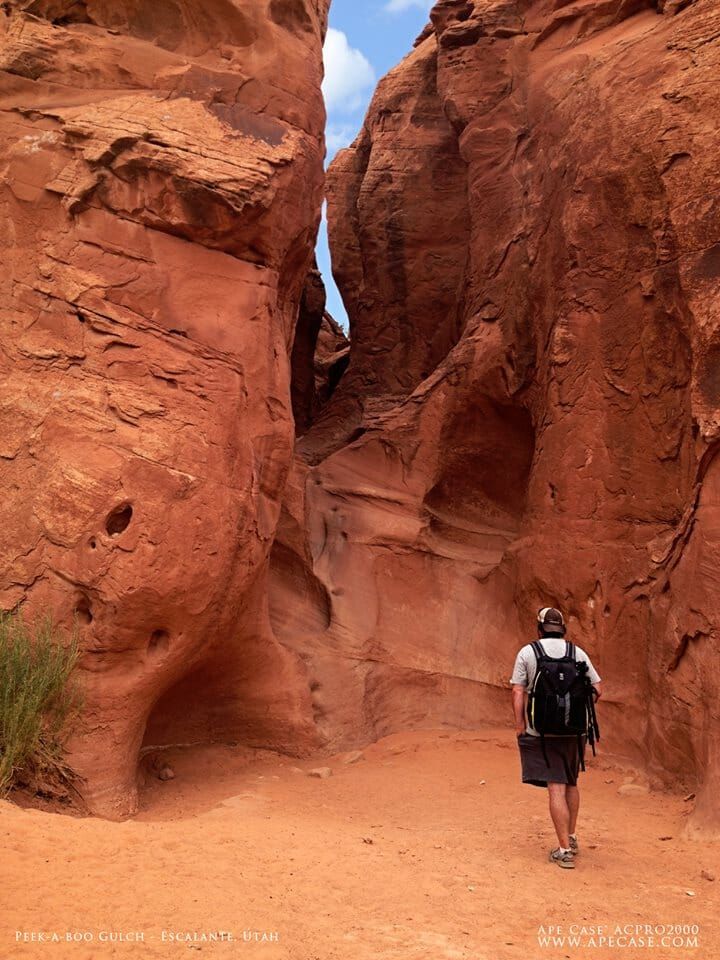 A hiker with a backpack walks toward a narrow opening between tall, textured red rock canyon walls under a blue sky.