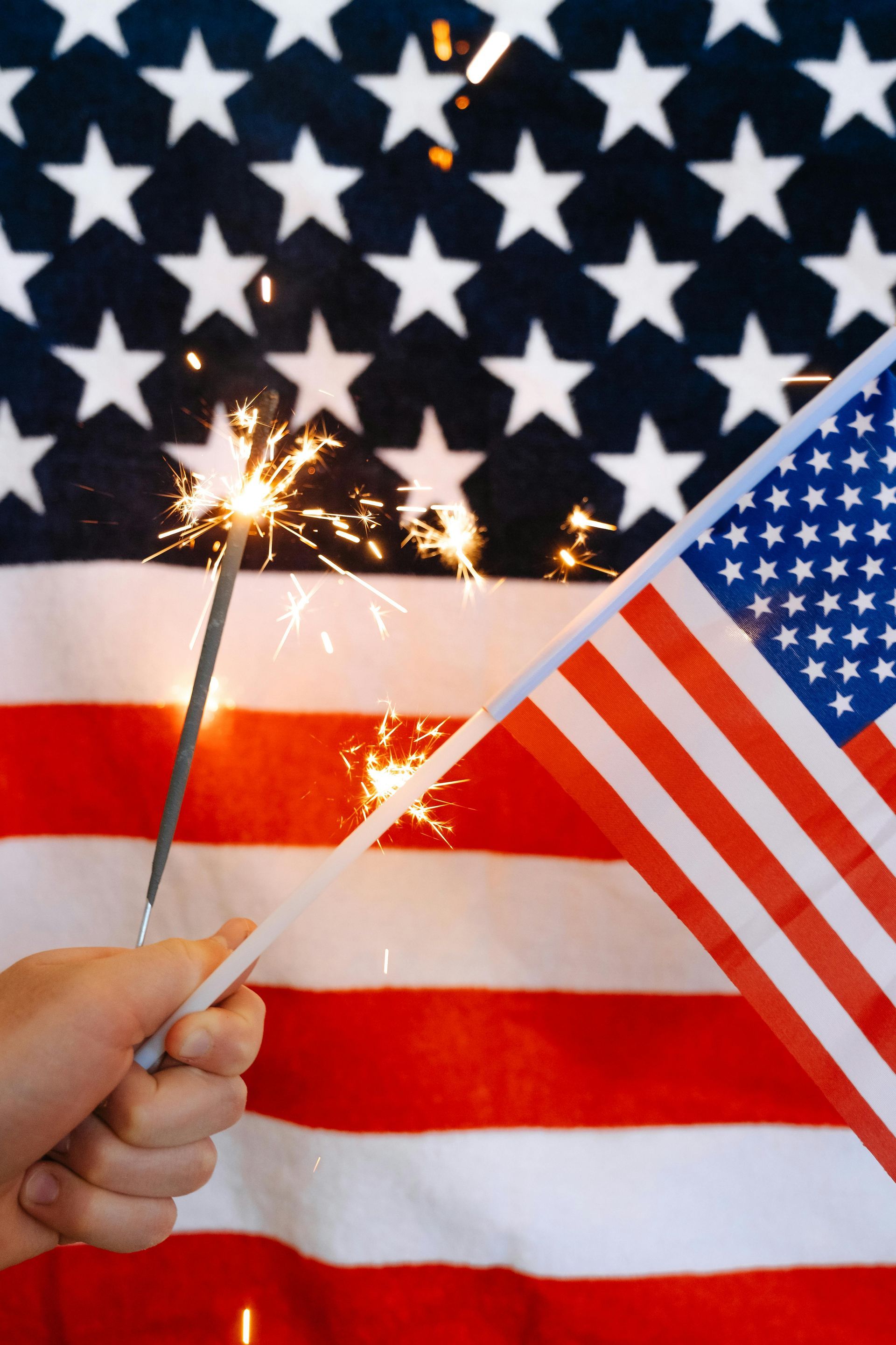 A hand holds a glowing sparkler and a small American flag in front of a larger American flag backdrop.