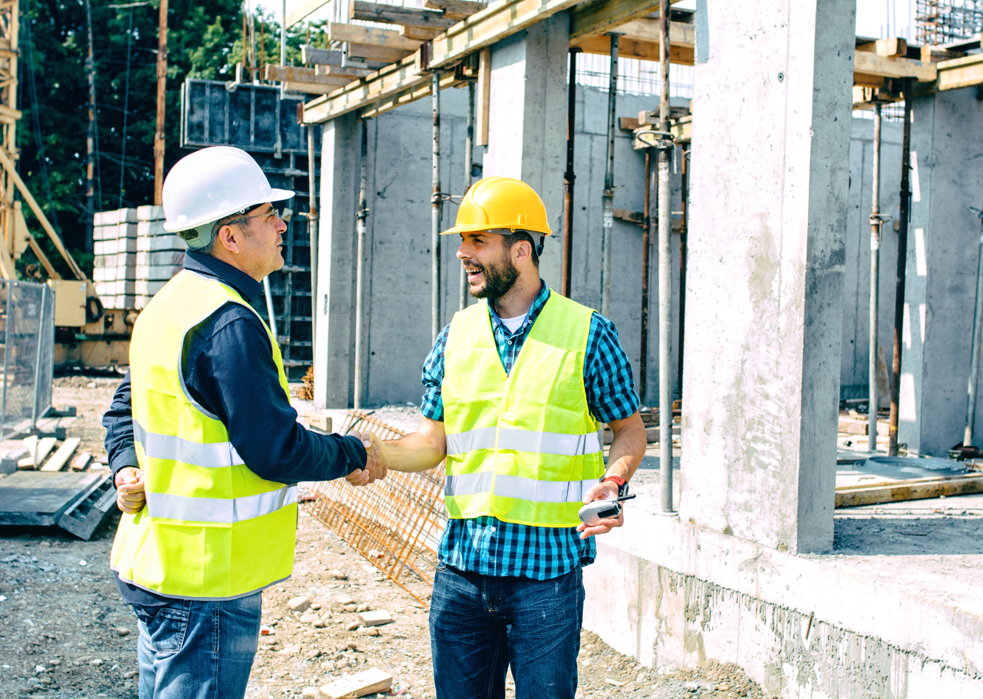Two construction workers in safety vests shake hands on a construction site.