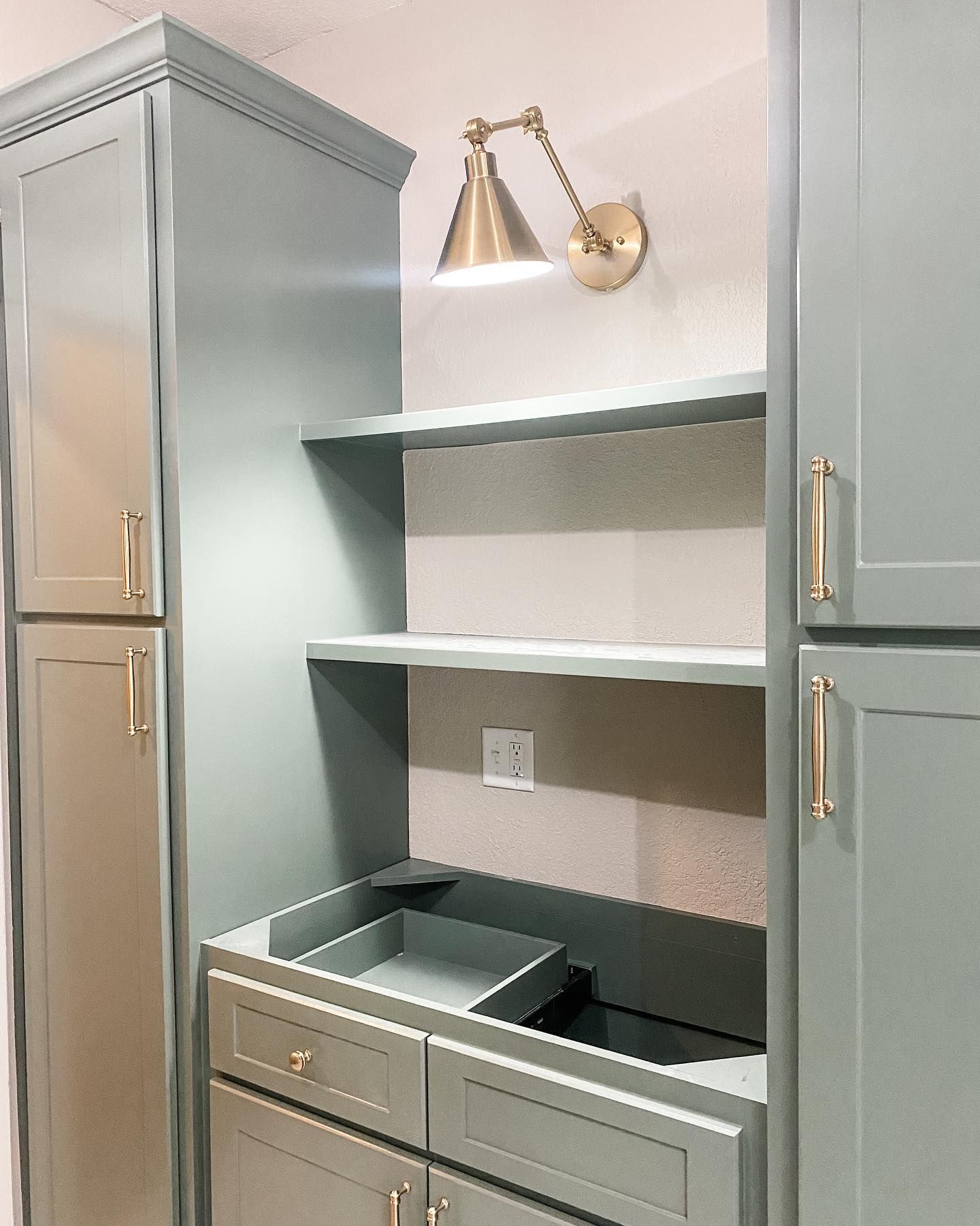 Built-in cabinet with open shelves, a desk area, and a gold sconce light. Painted a soft green/gray.
