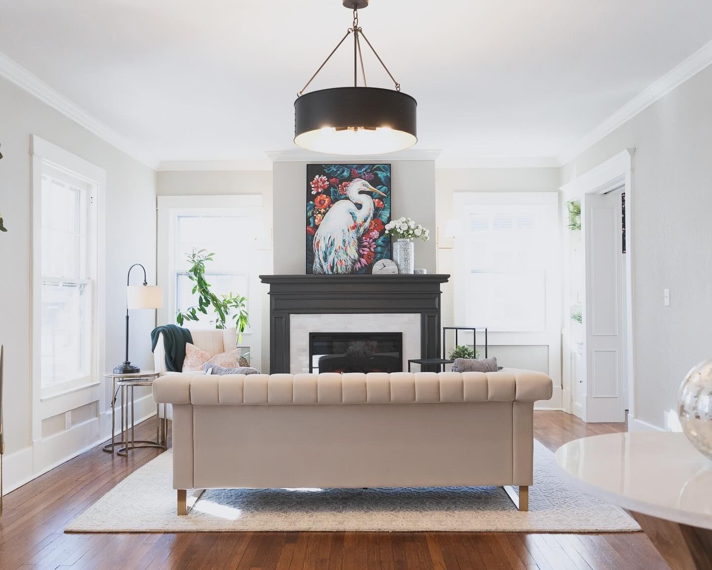 Living room with neutral color scheme, featuring a sofa, fireplace with artwork, and dark chandelier.