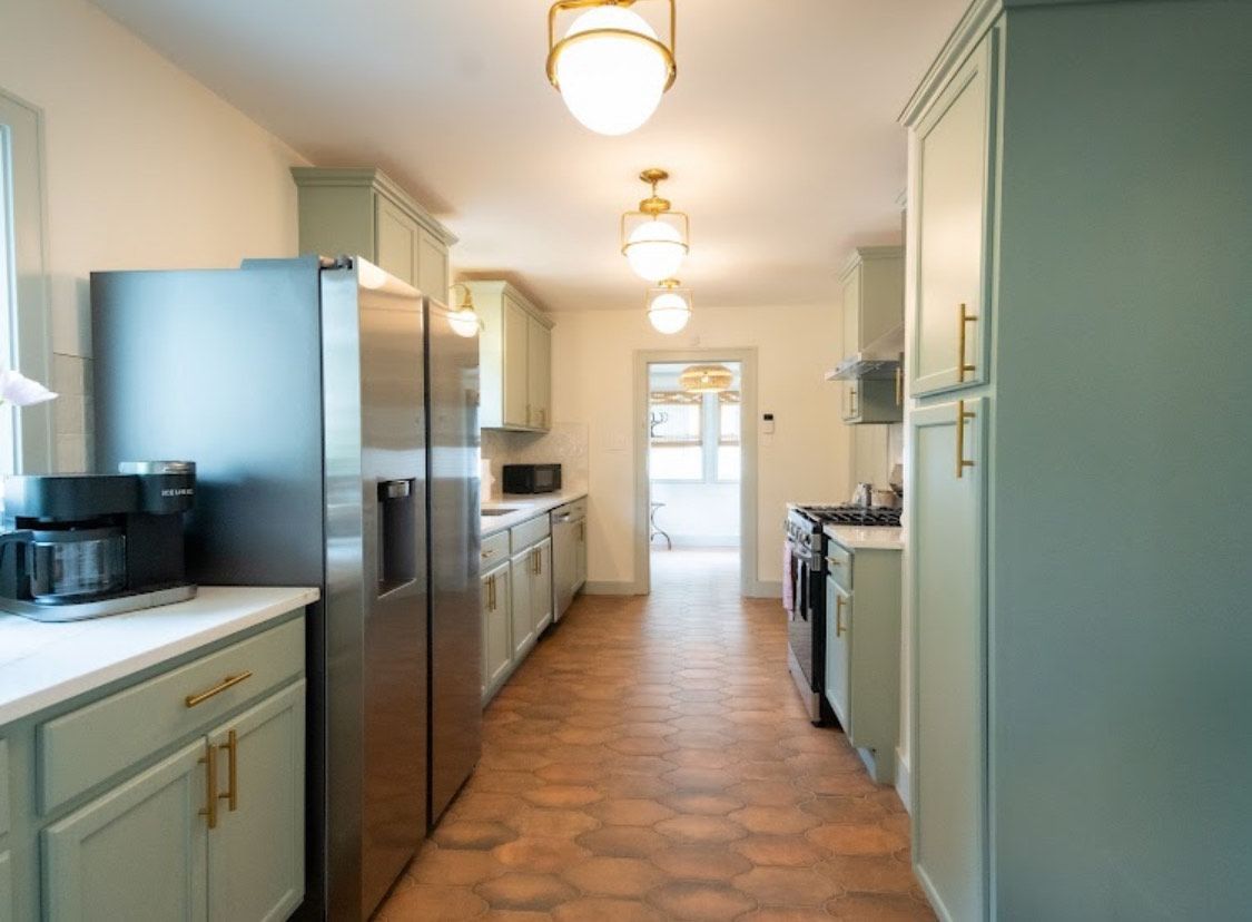 A long, narrow kitchen with sage green cabinets, stainless steel appliances, and patterned terracotta floor.
