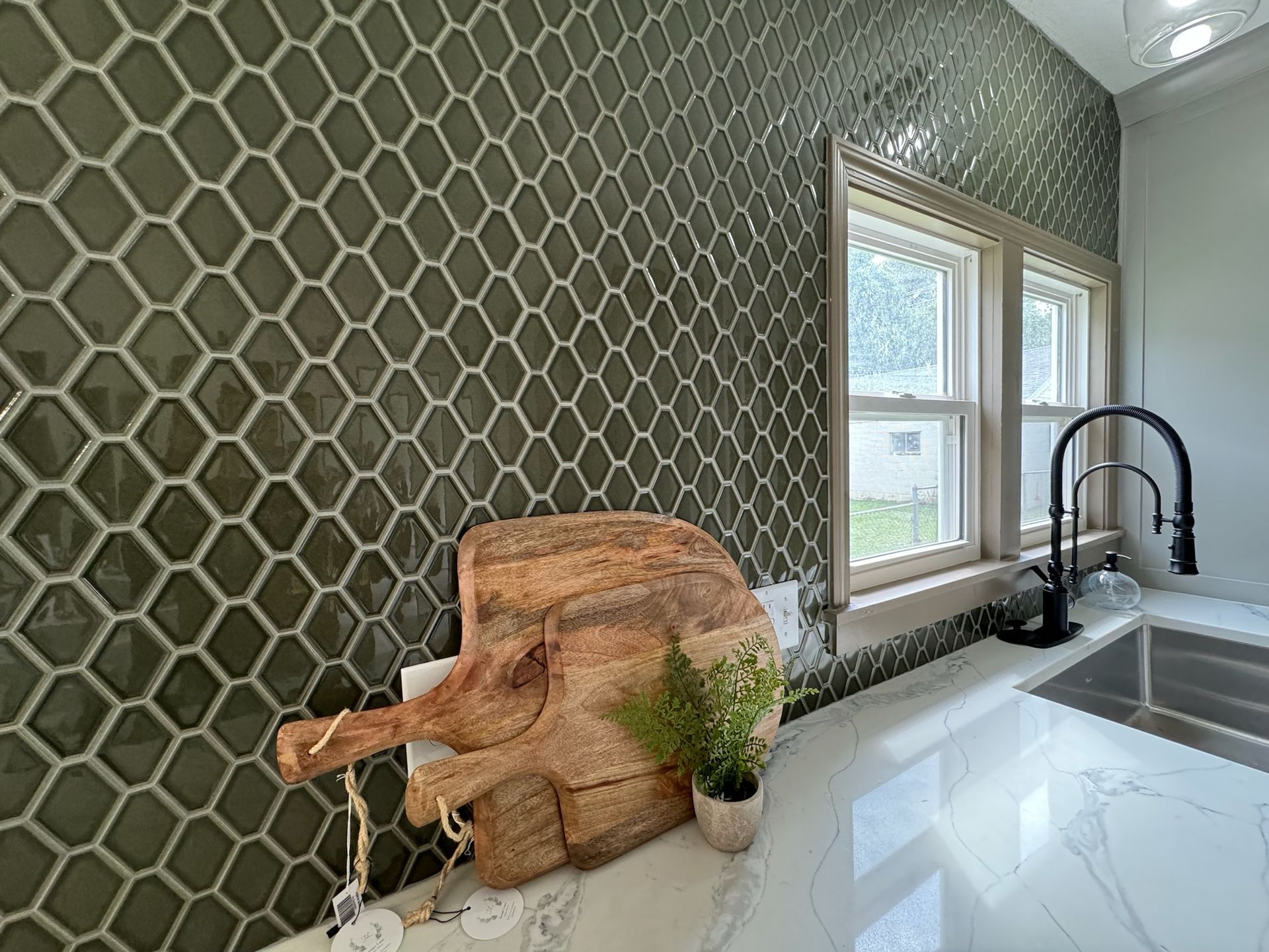 Kitchen with olive green patterned tile backsplash, white countertops, and two wooden cutting boards.