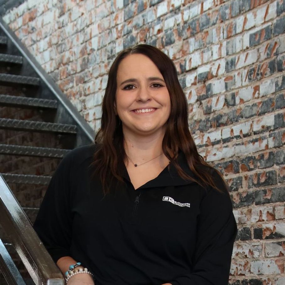 Woman with long brown hair smiles, leaning on a stair rail, against a brick wall.