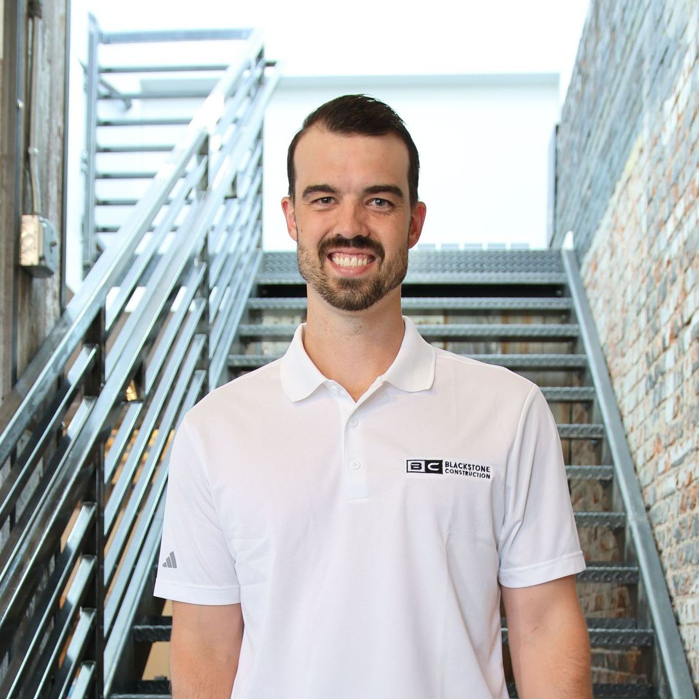 Man in white polo smiling in front of a staircase, brick wall on right, wooden wall on left.