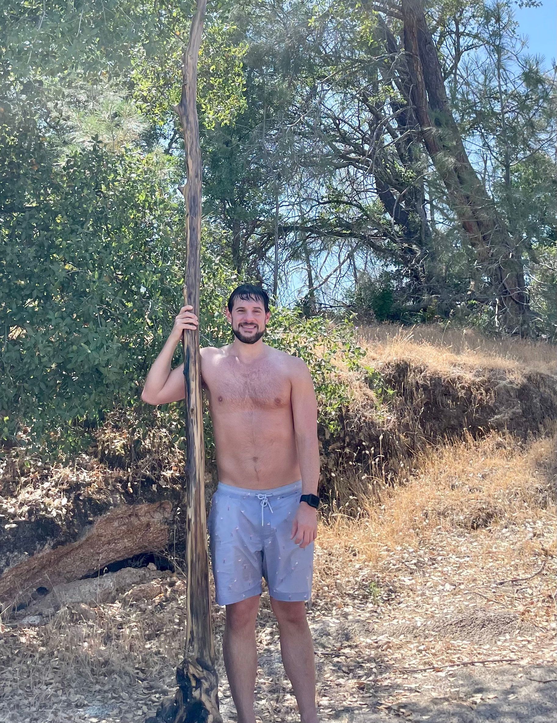 A shirtless man is standing in the dirt holding a long stick.