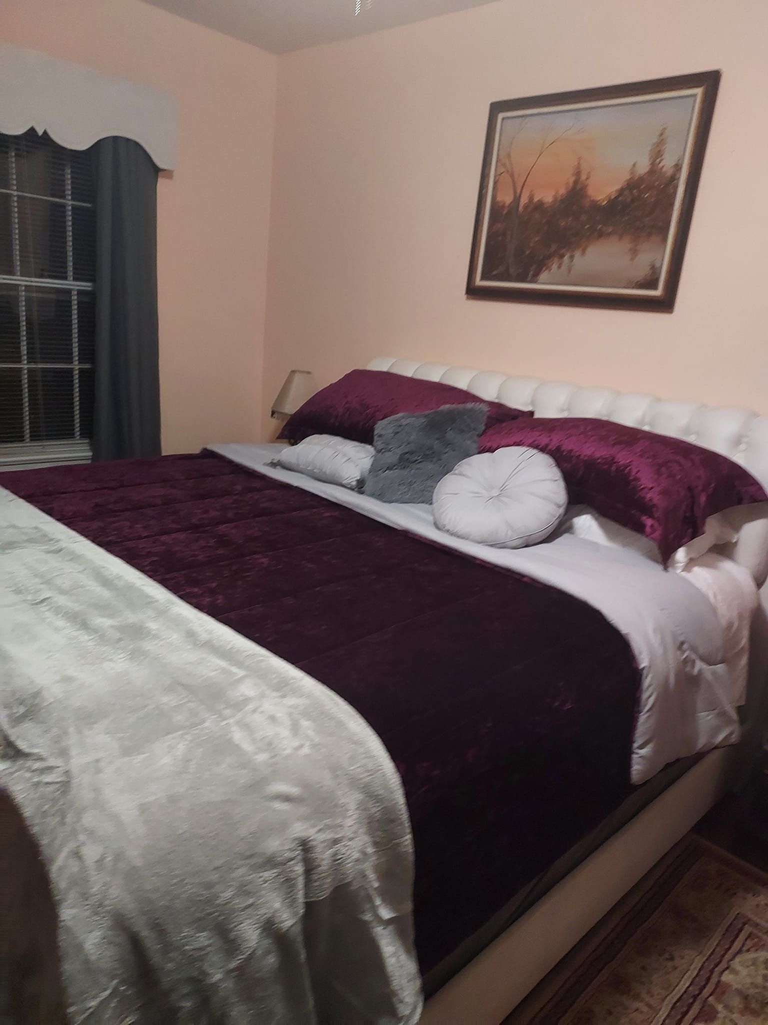 Cozy bedroom with a large bed. Deep purple and grey bedding, framed painting, and window with dark curtain.