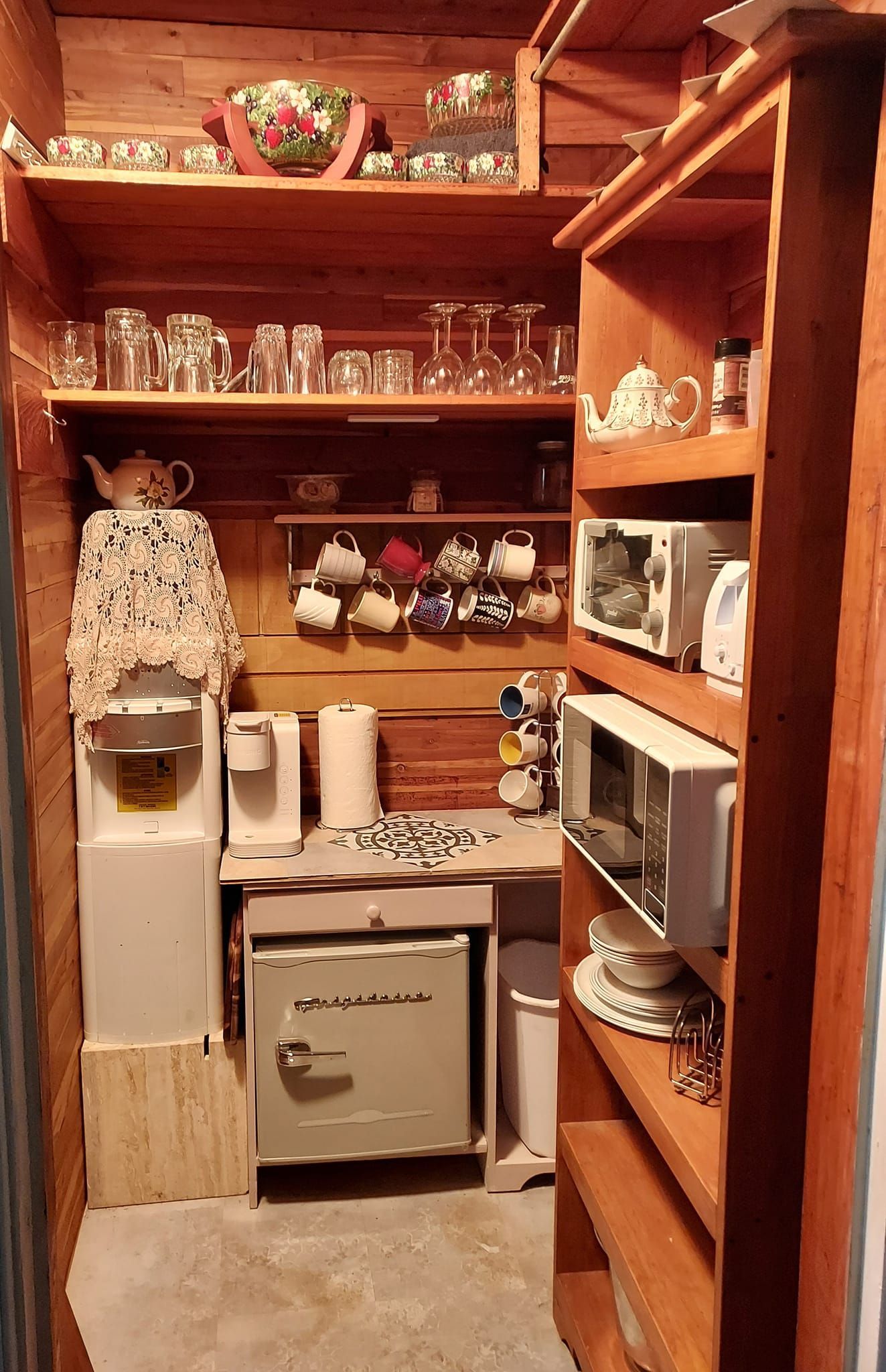 Small rustic kitchenette with wooden shelves, containing dishes, mugs, and appliances like a microwave and refrigerator.