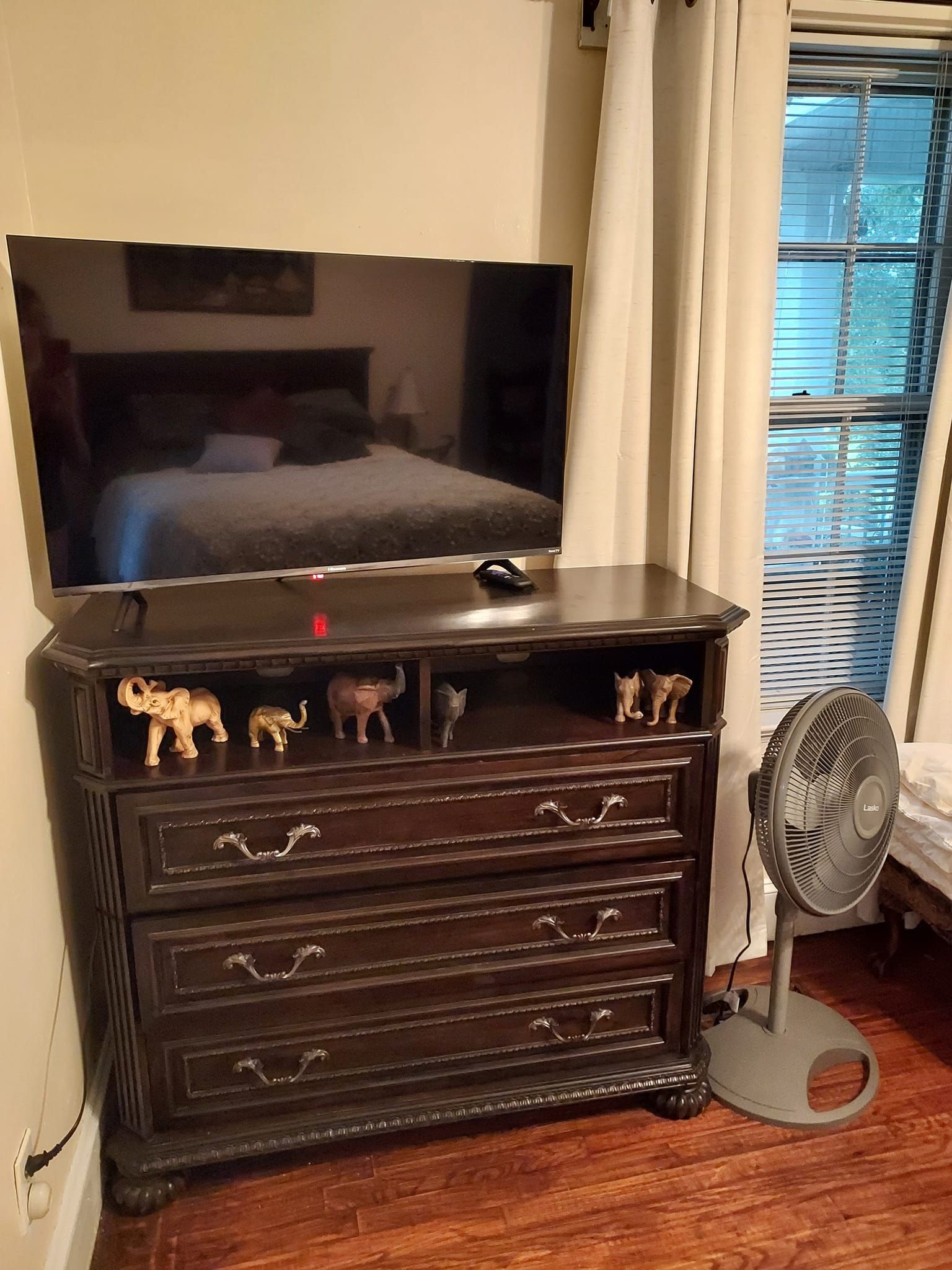 TV on a dark wood dresser with animal figurines, next to a window and a fan.
