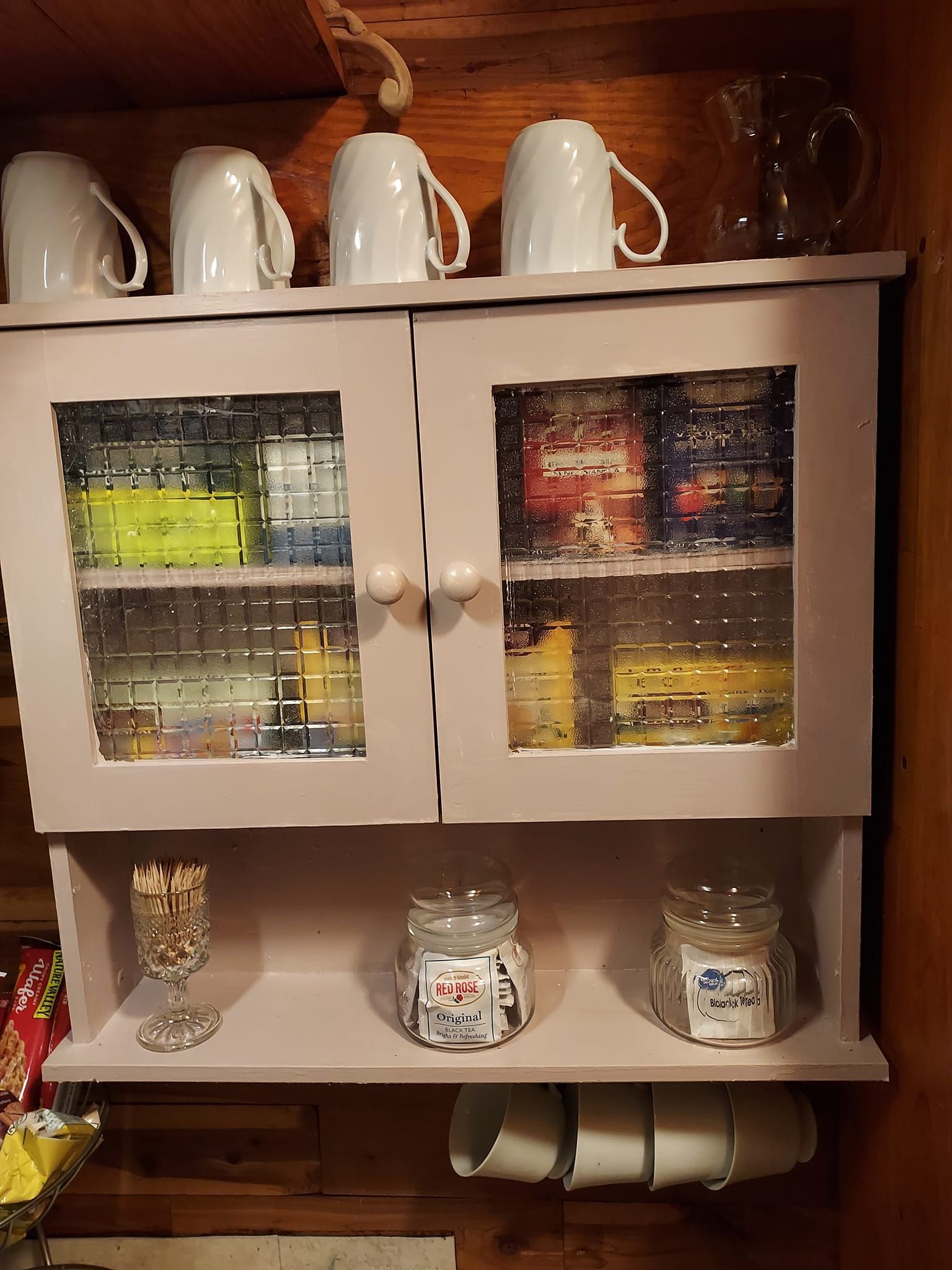 A light purple cabinet with glass doors holding tea, mugs above, jars below.