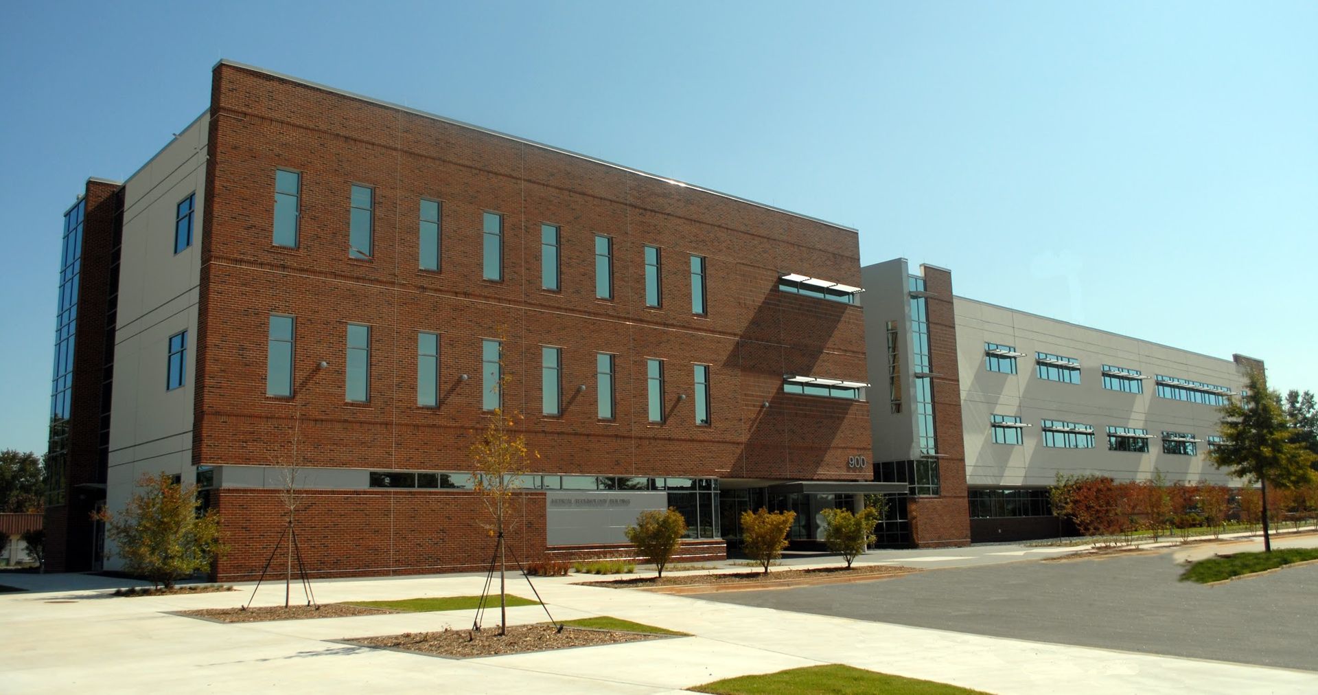 Three-story brick building with arched windows, light-colored side, small trees, and a clear blue sky.