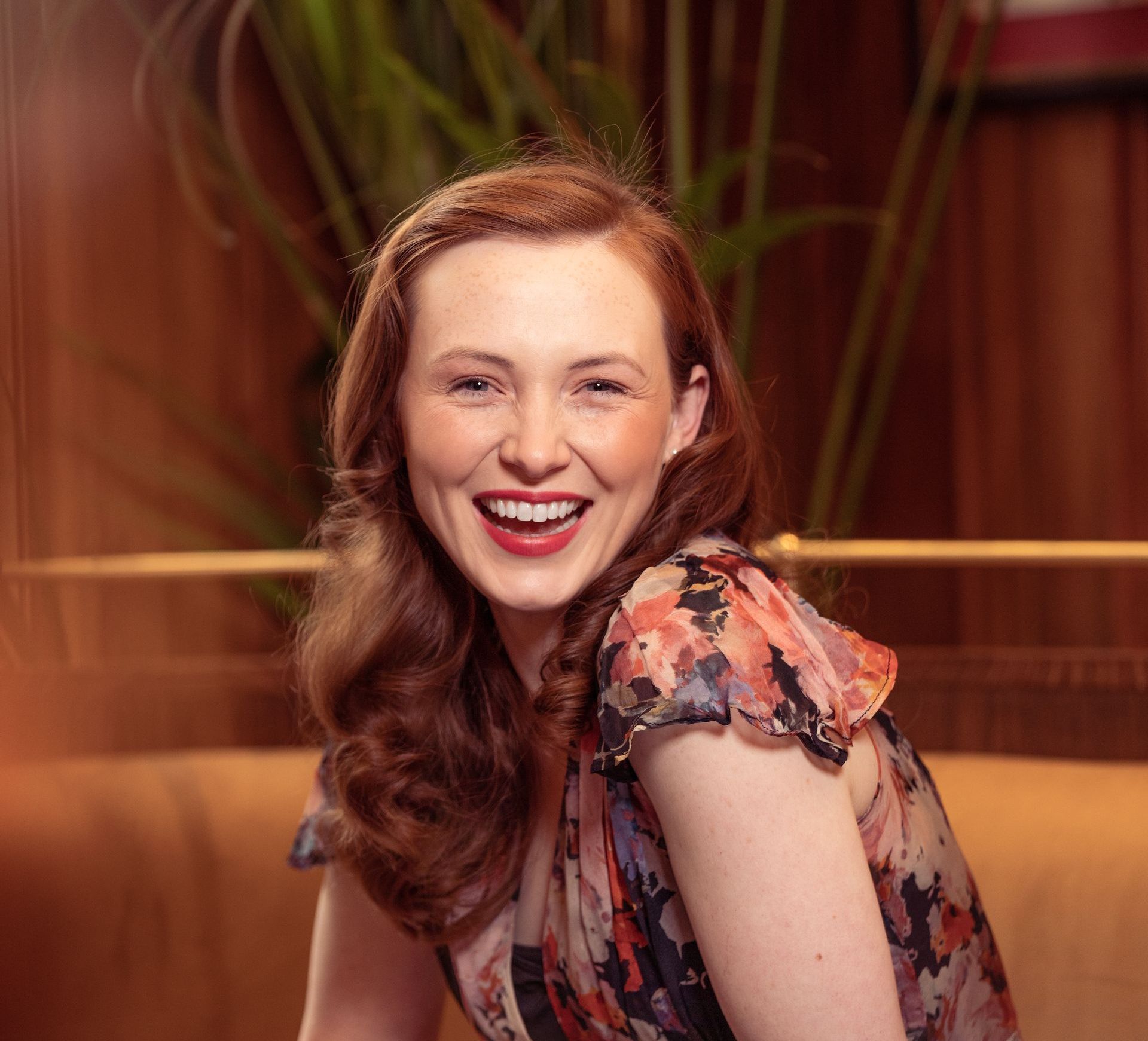 A woman in a floral dress is smiling and sitting on a couch.