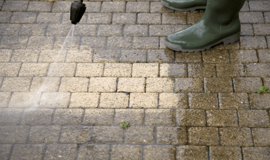A person wearing green boots power washes a brick patio, cleaning the surface.