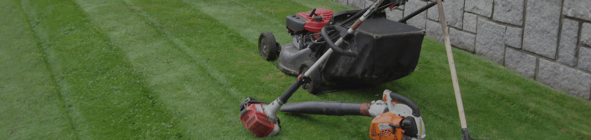 Lawn mower, weed wacker, and a leaf blower on freshly cut green grass next to a stone wall.