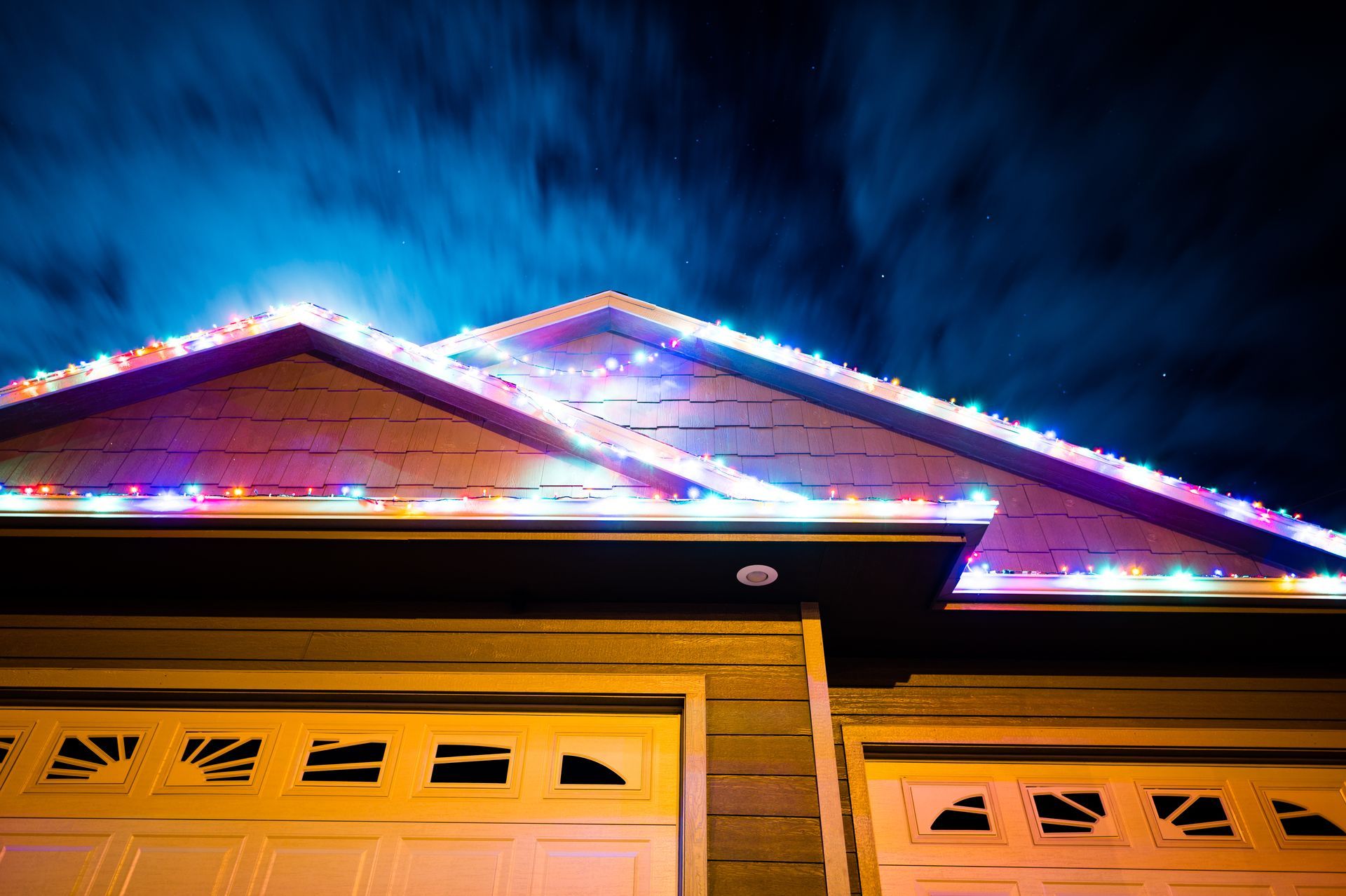 House with Christmas lights on roof, garage doors below. Lights glow against a dark, cloudy night sky.
