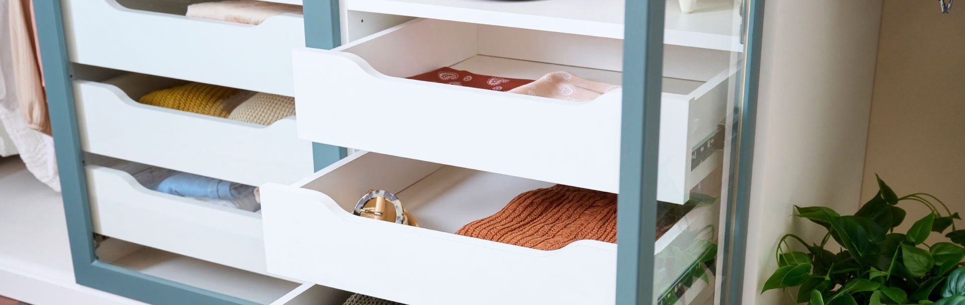 A dresser with three drawers filled with clothes and a plant in the background.