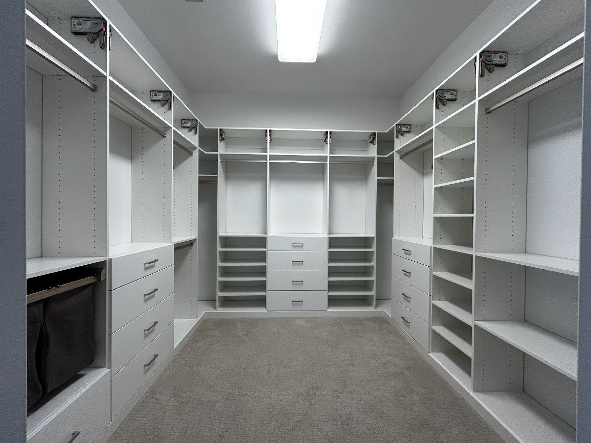 Empty, white walk-in closet with shelves, drawers, and hanging rods. Gray carpet. Bright overhead lighting.