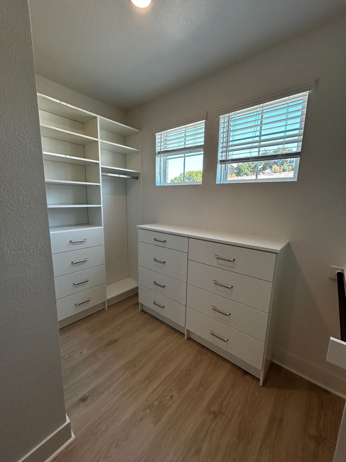 A white walk-in closet with shelves, drawers, hanging space, and two windows.  Light wooden floor.