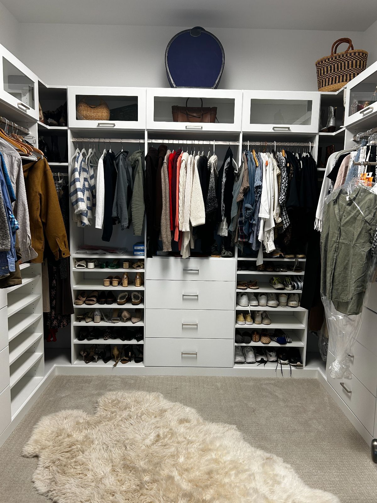 Walk-in closet with white shelves, drawers, and rods holding clothes. A faux fur rug lies on the floor.
