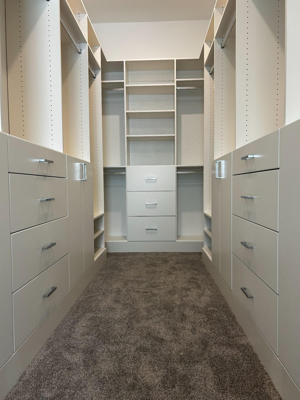 A well-lit walk-in closet with white cabinets, drawers, and shelving. Dark gray carpeted floor.