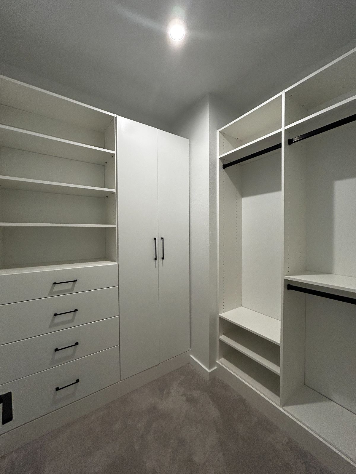 White walk-in closet with shelves, drawers, and hanging rods. Gray carpet, neutral walls, and overhead lighting.
