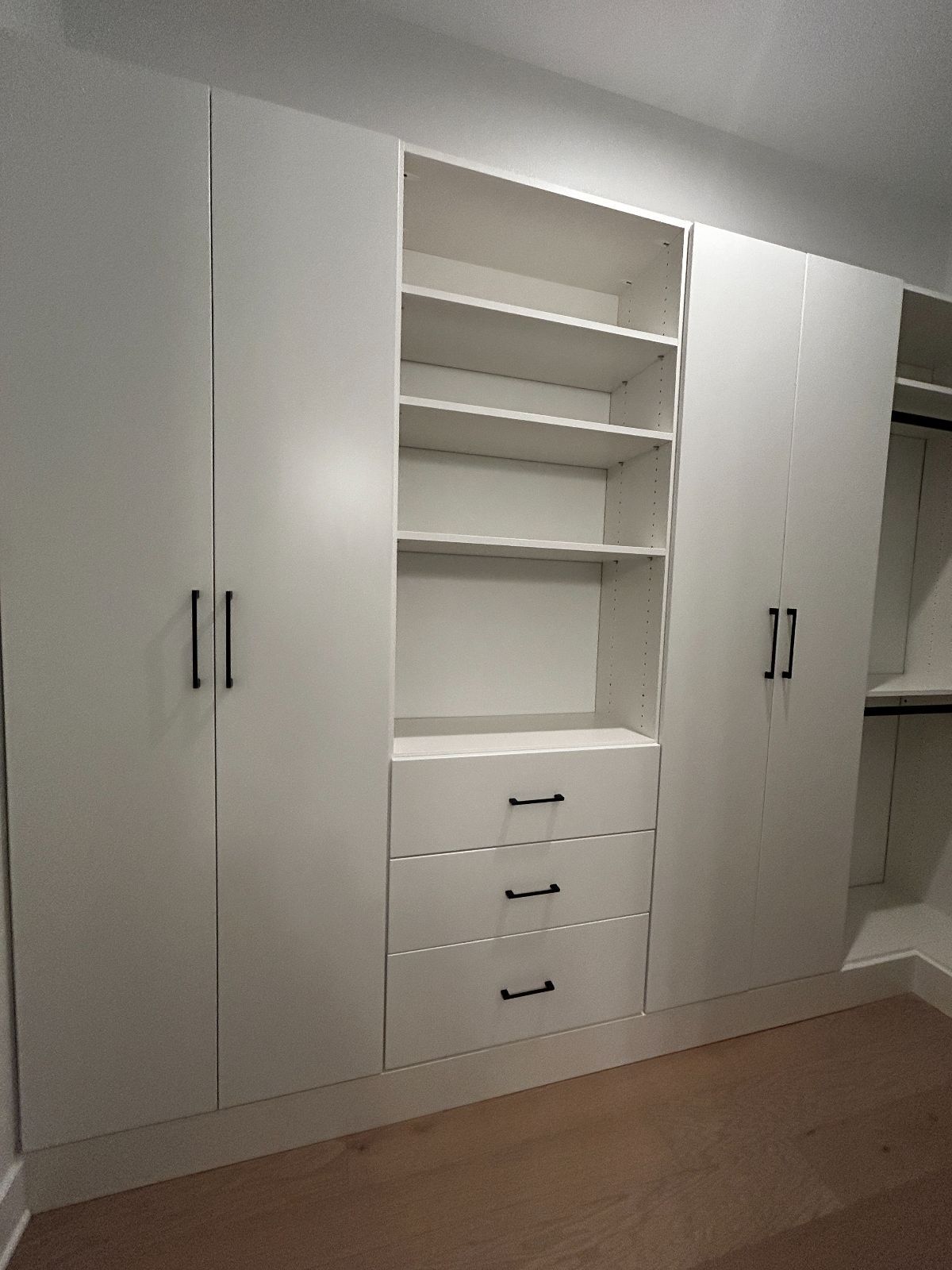 White built-in closet with shelves, drawers, and doors, against a white wall and hardwood floor.