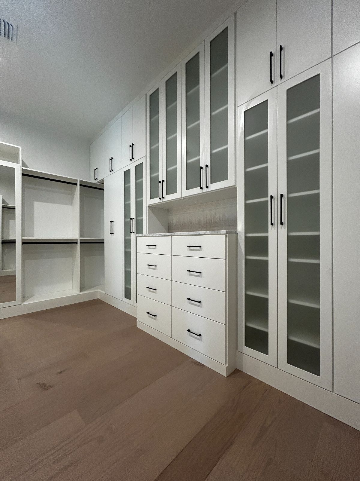Walk-in closet with white cabinets, frosted glass doors, and light brown hardwood floors.
