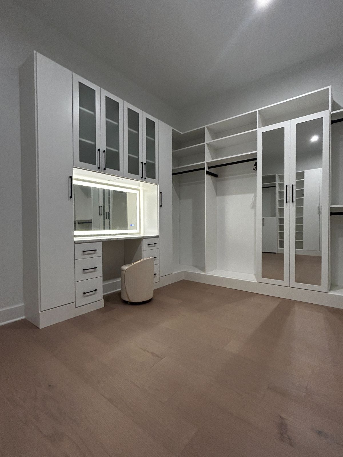 White walk-in closet with vanity, mirrored doors, and shelving against light wood flooring.