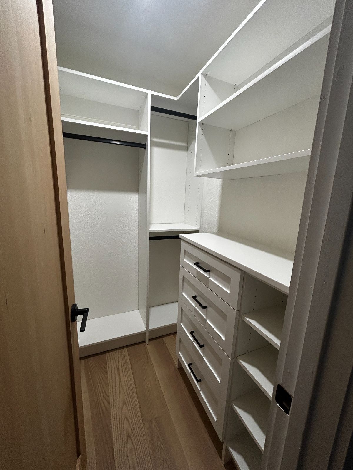 Walk-in closet with white shelves and drawers, dark rods and handles, light wood floor, and tan door.