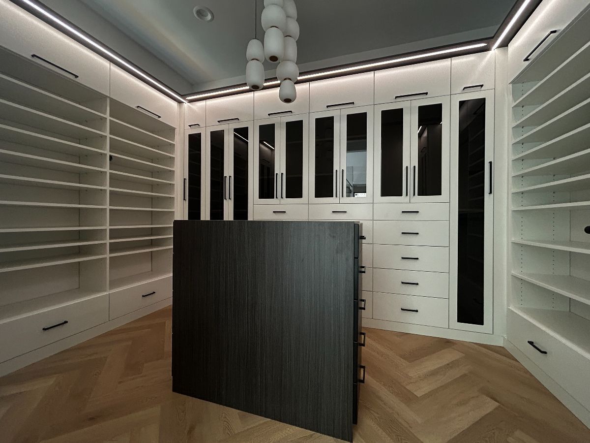 Walk-in closet with white shelves and cabinetry, central dark cabinet, and herringbone wood floor.