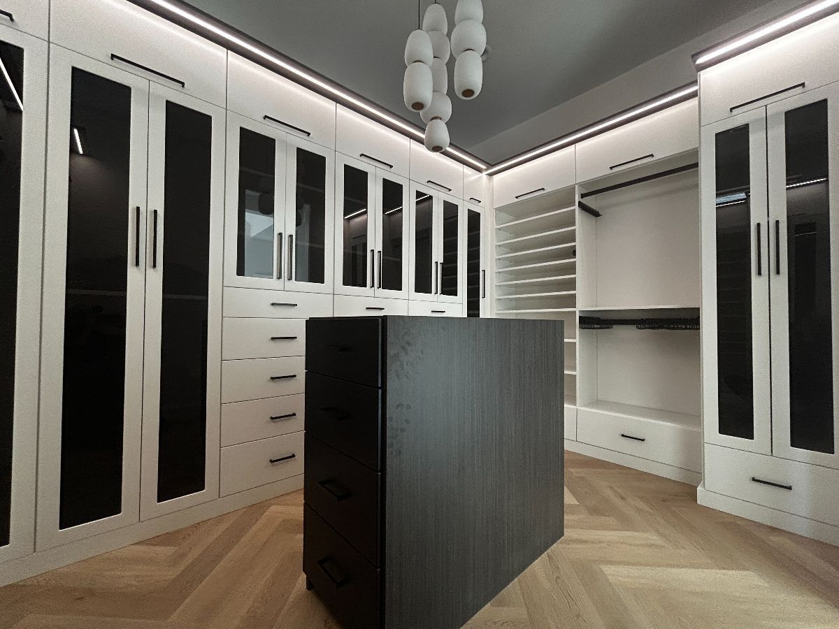 Walk-in closet with white cabinets, dark glass doors, a central dark wood island, and overhead lighting.