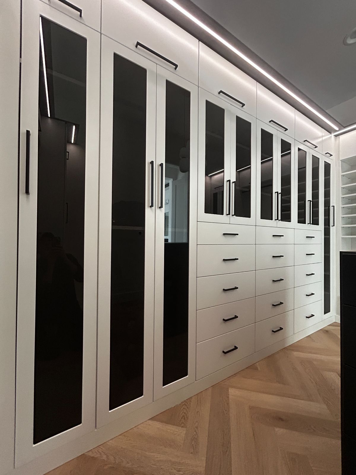 White custom closet with glass doors, black handles, and drawers. Wooden chevron floor.