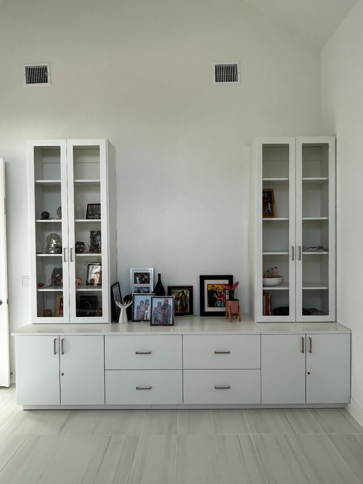 White built-in cabinet unit with glass-door upper cabinets, lower storage cabinets, drawers, and display shelf against a white wall.