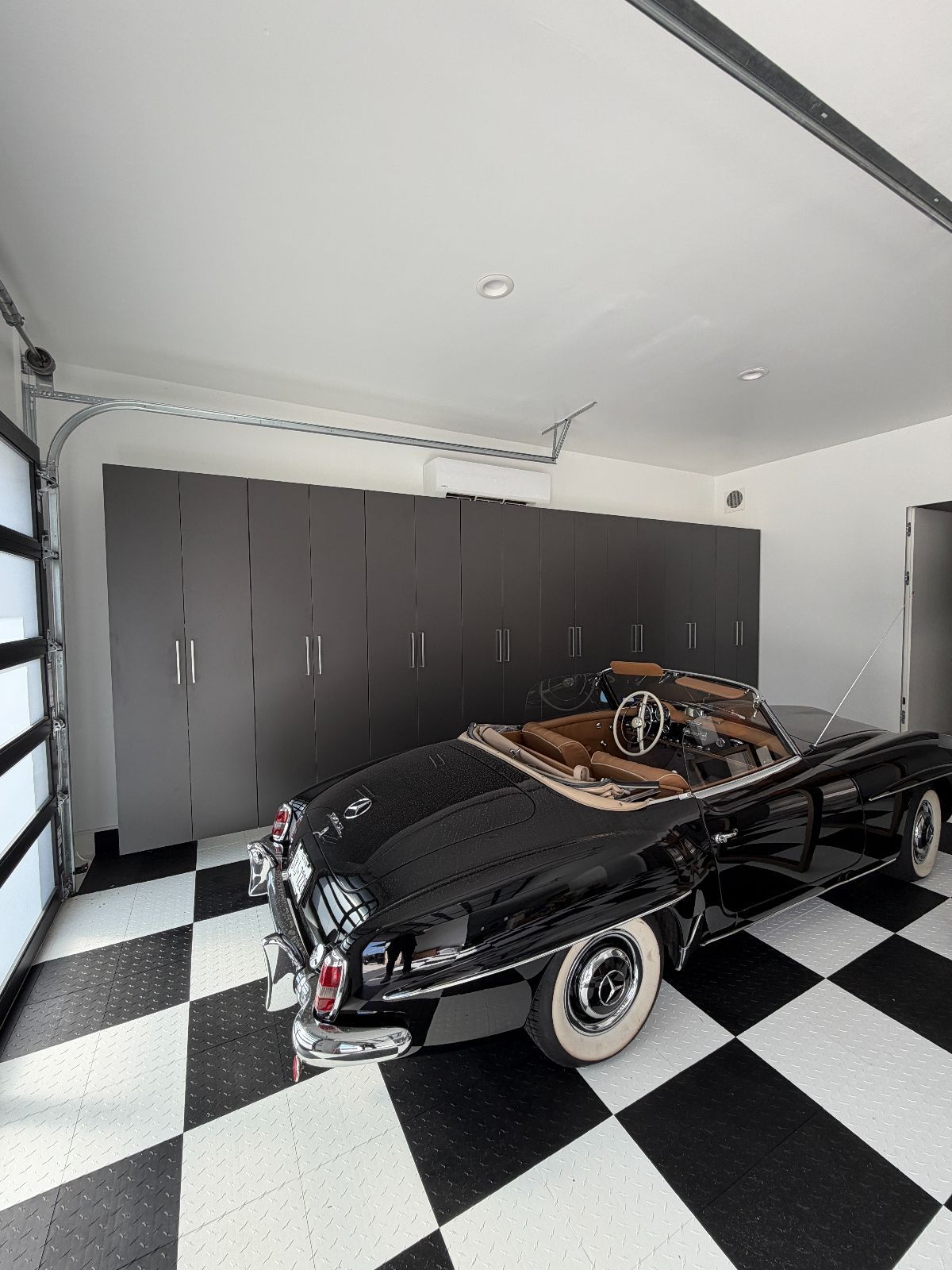 Black vintage car in a garage with black and white checkered floor, cabinets, and a glass garage door.