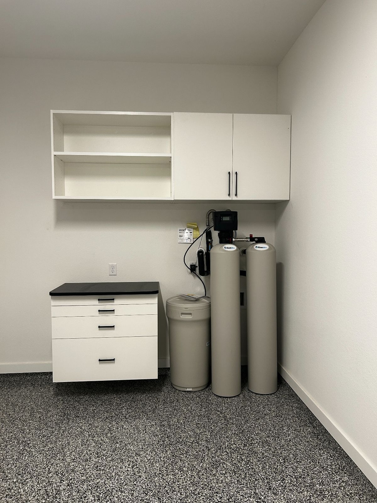 White cabinets, storage, and water treatment system in a utility room with speckled flooring.