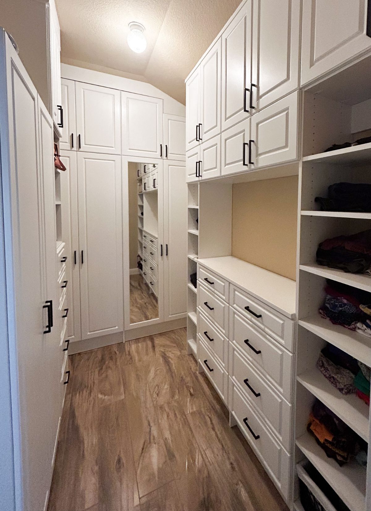 A walk in closet with white cabinets , drawers , shelves and a mirror.