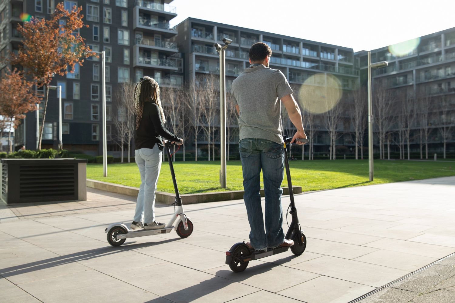 A man and a woman are riding electric scooters down a sidewalk.