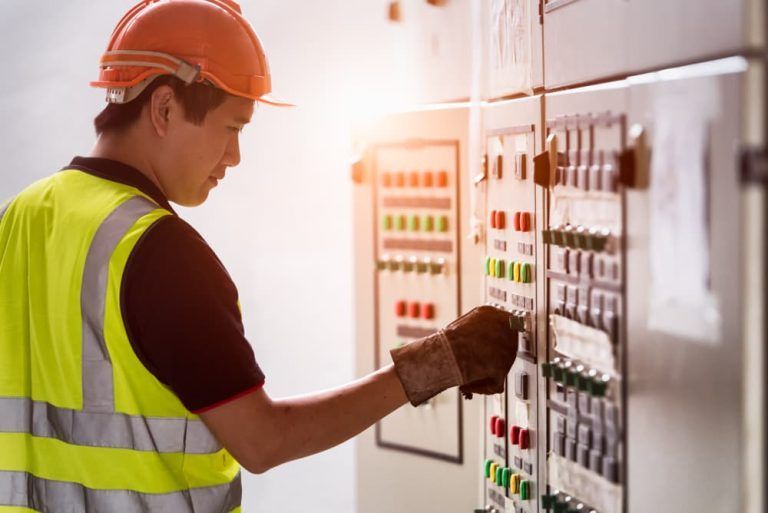 A man wearing a hard hat and safety vest is working on a control panel.
