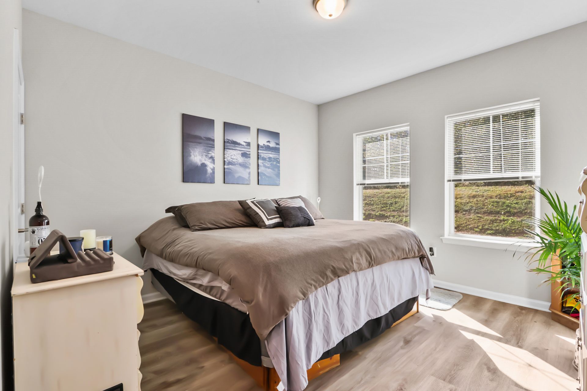 Bedroom with a bed, dresser, windows, and wall art. Gray walls, brown bedding, and wood-look flooring.