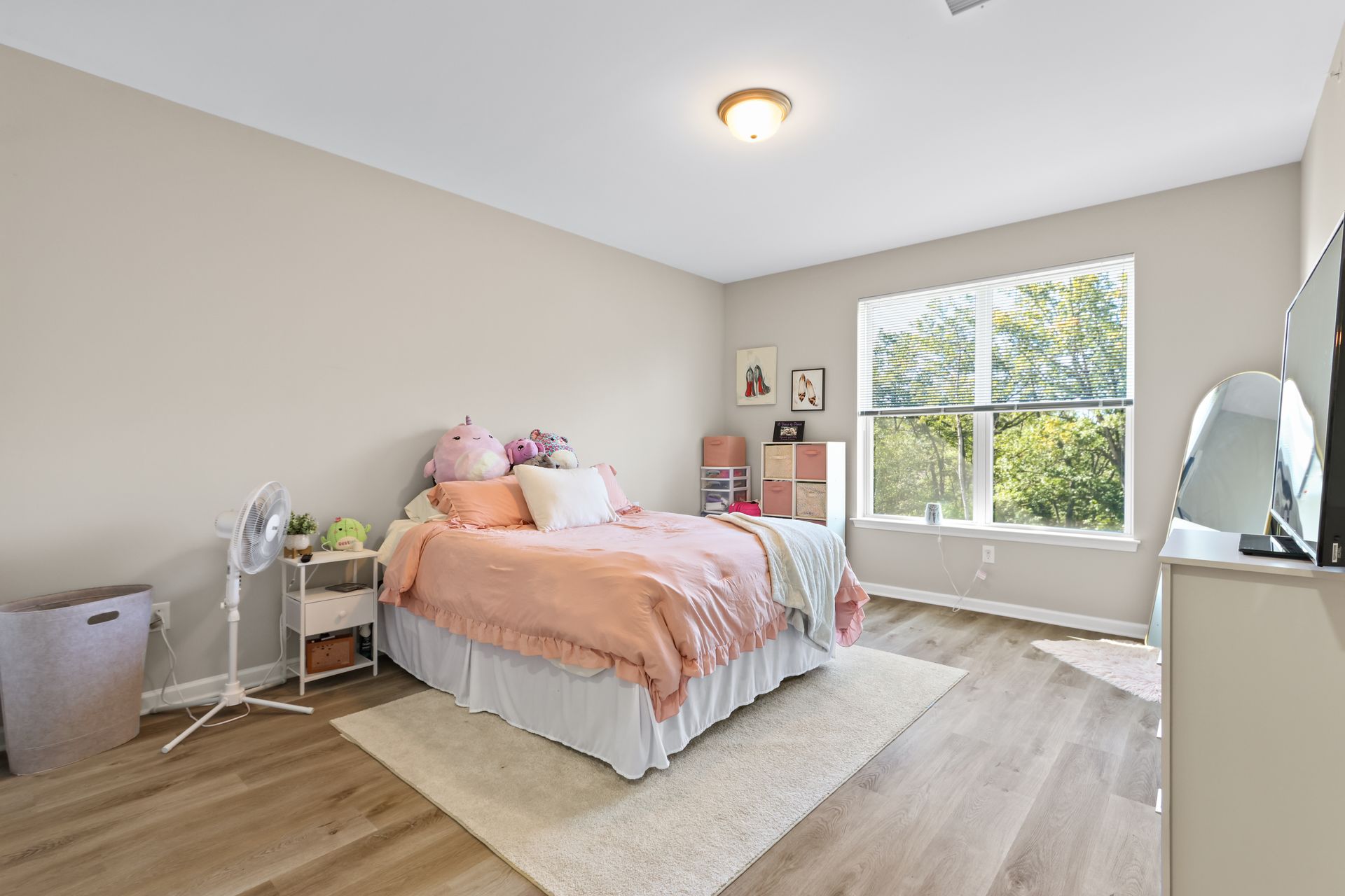 Bedroom with a bed, dresser, window, and fan. Pink and white decor, neutral walls, light wood floor.