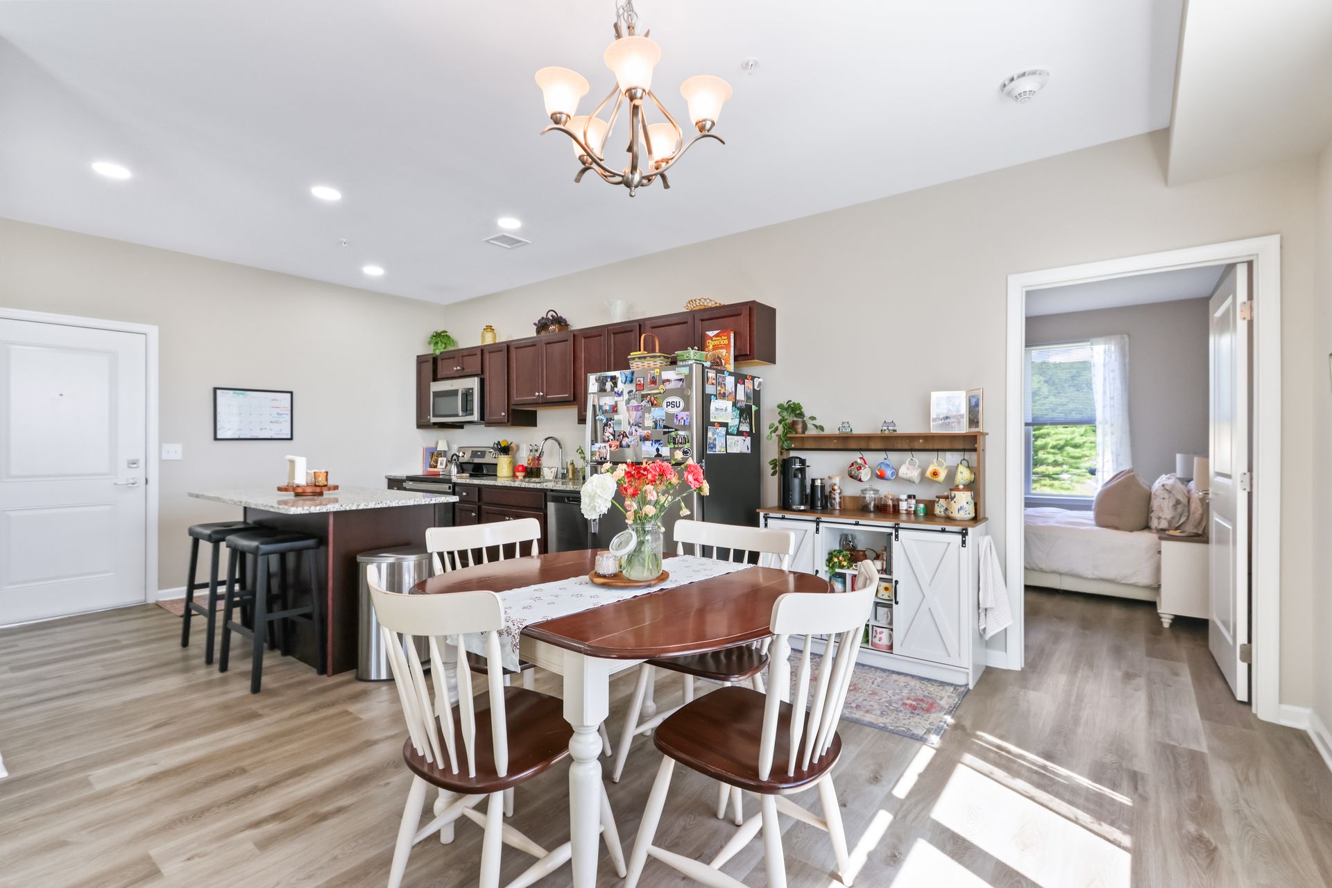 Kitchen and dining area with dark cabinets, a table, and chairs. Bedroom visible in the background.