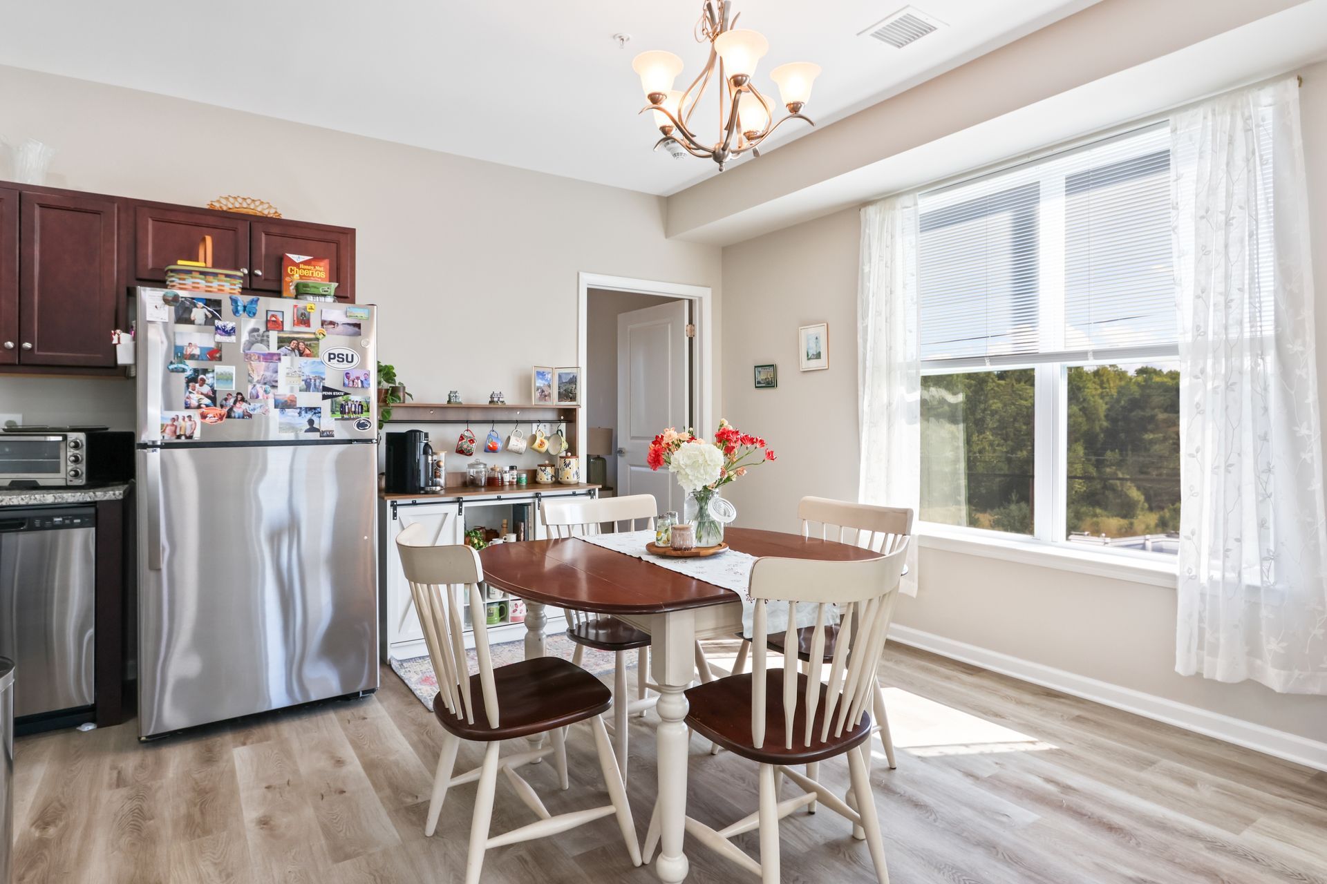 Kitchen with stainless steel refrigerator, wooden dining table with four chairs, and large window with sheer curtain.