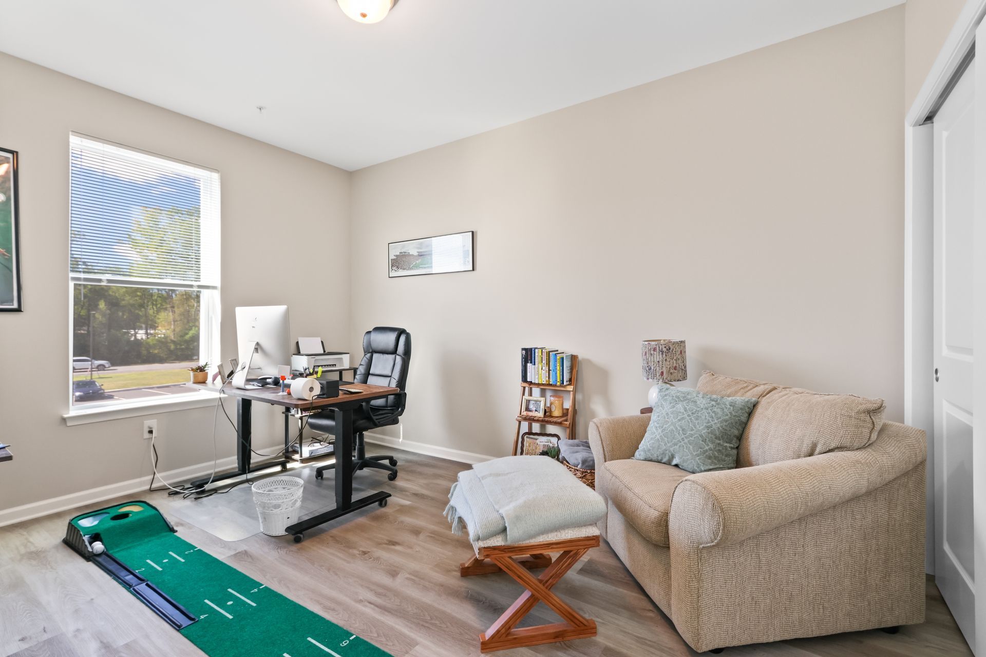 Home office with desk, chair, couch, and putting green. Neutral tones, natural light.