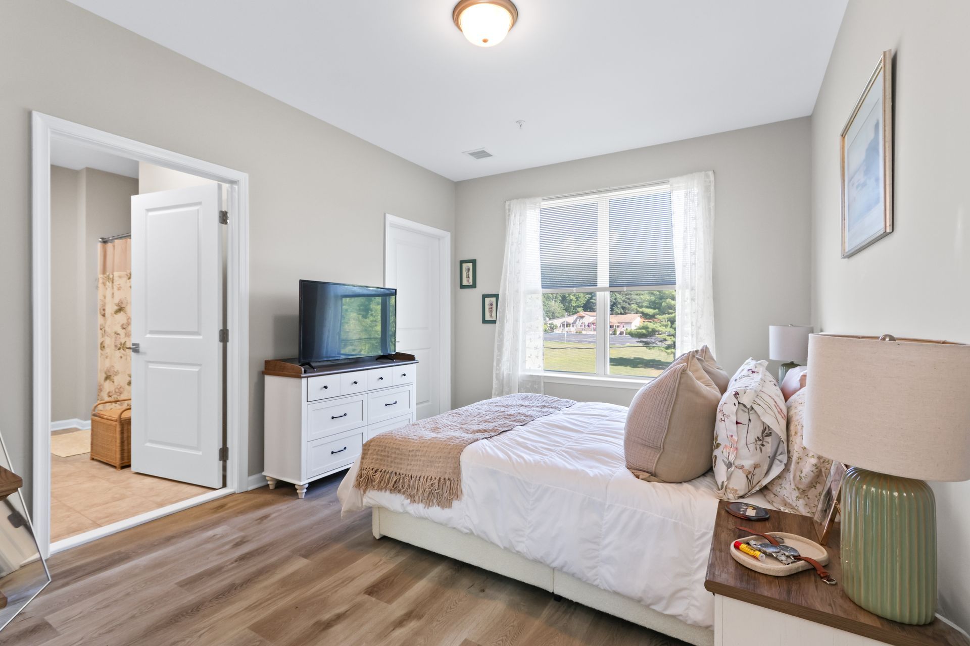 Bedroom with white bed, dresser, TV, open doorway to bathroom, and window with view.