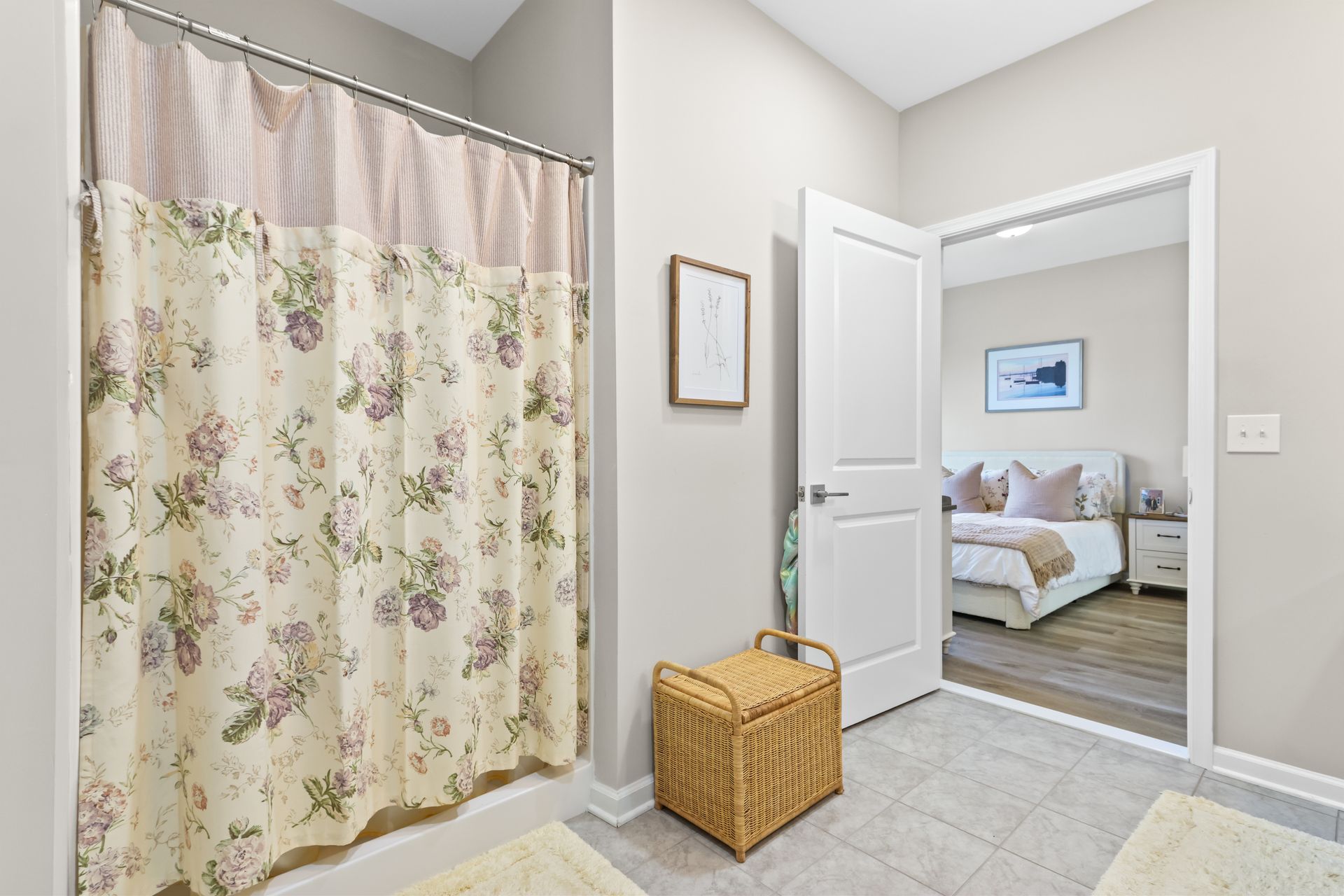 Bathroom with floral shower curtain, open door to bedroom with bed, beige walls, wicker basket.
