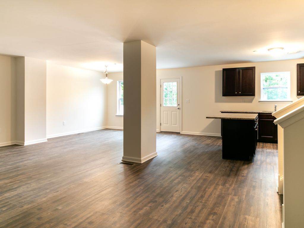 Open-concept living space with dark wood-look floors, white walls, kitchen with dark cabinets, and a door to the outside.