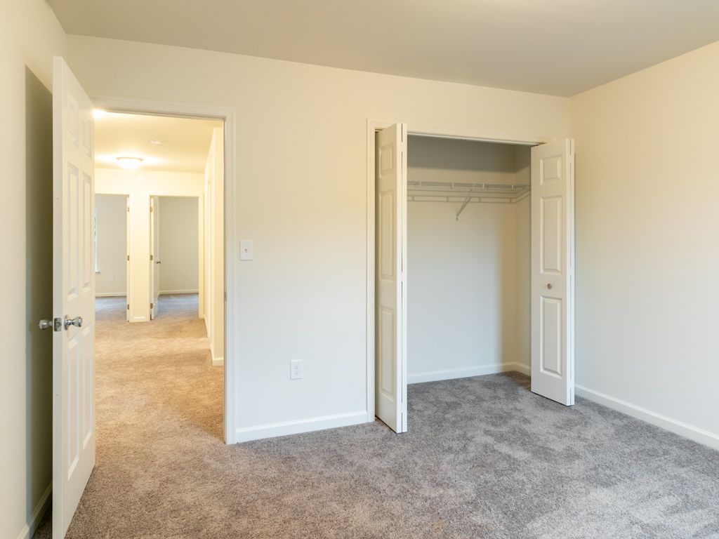 Empty bedroom with closet and doorway to hallway; gray carpet, white walls.