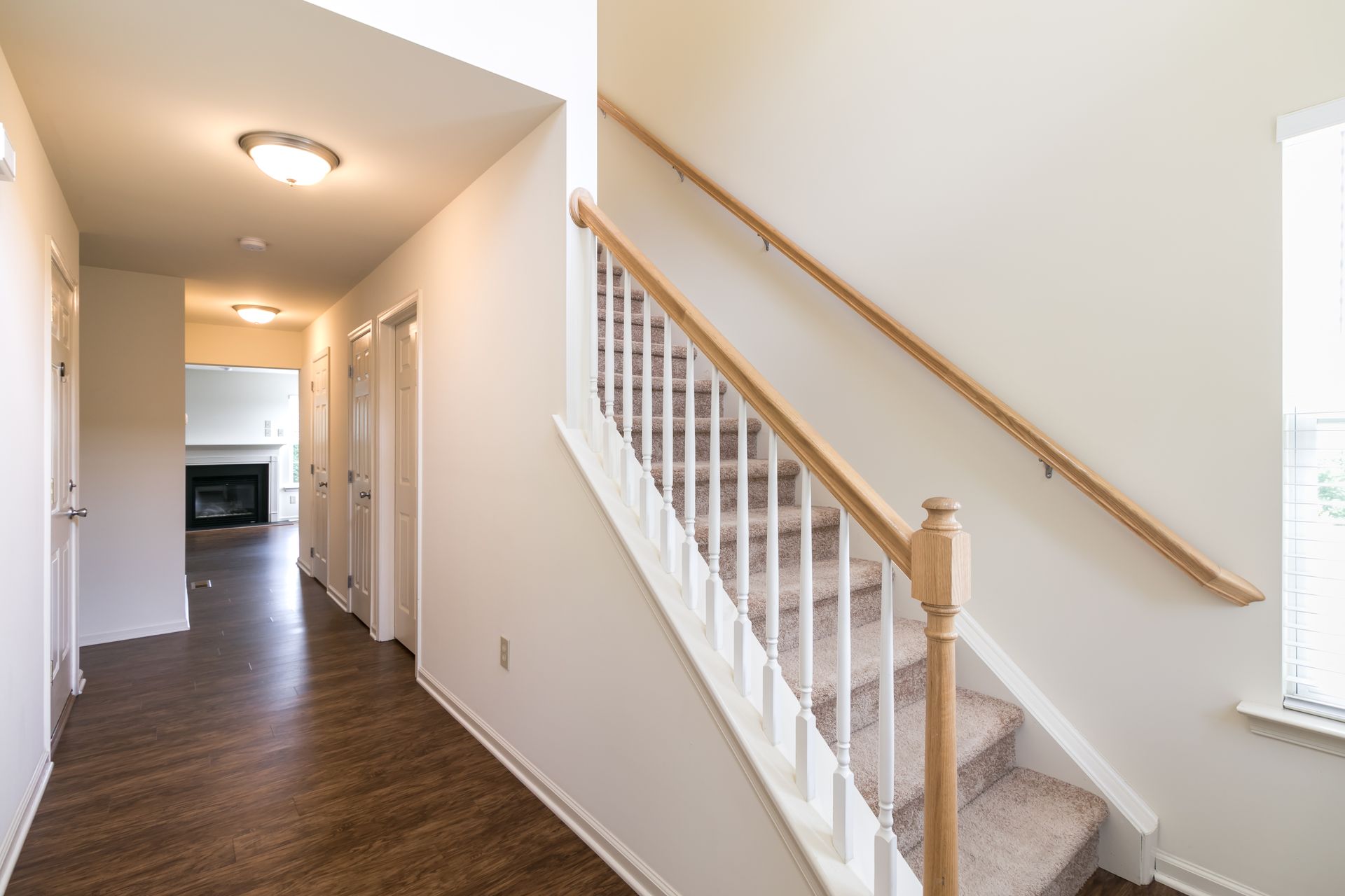 Hallway with staircase, tan carpet, and white walls leads to a living room with a fireplace.