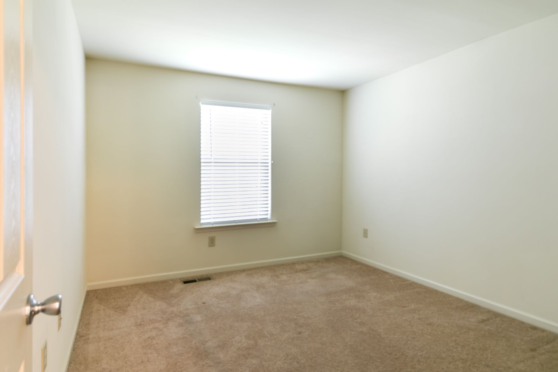 Empty bedroom with beige carpet, window with blinds, and light-colored walls.